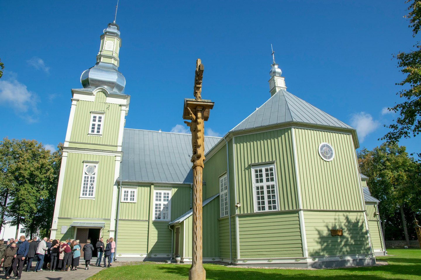People gather outside a light green wooden church with a steeple.